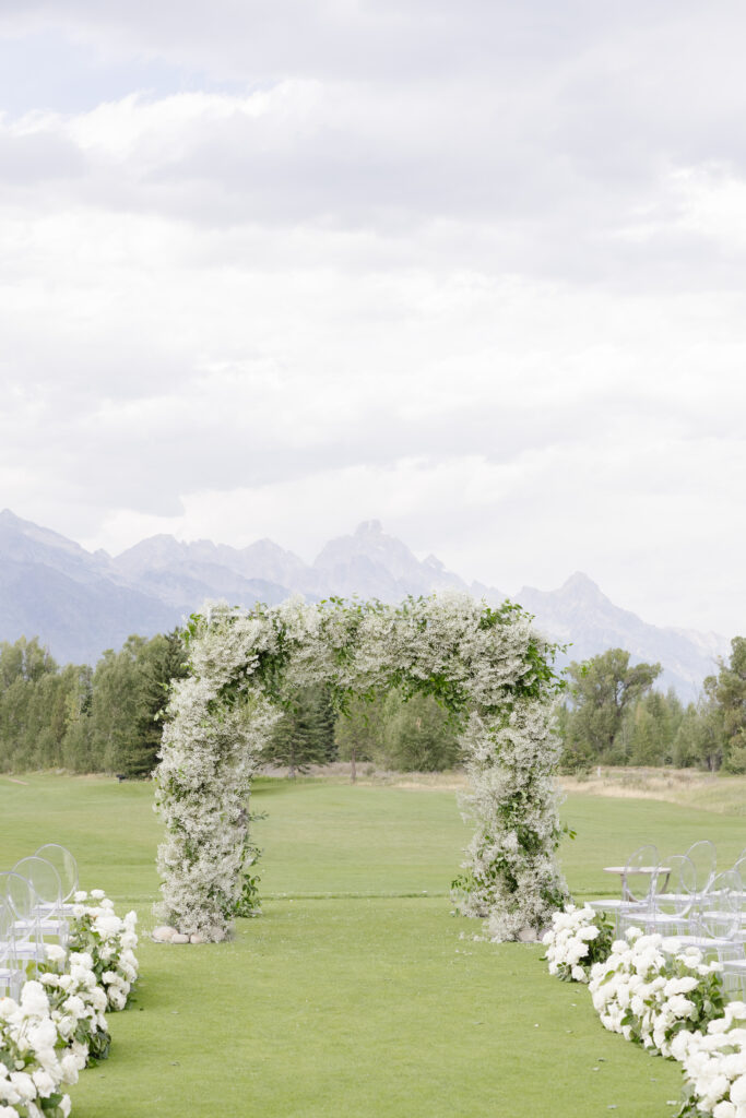 Elegant ceremony setup at Jackson Hole Golf and Tennis Club with white roses and ghost chairs. You said: ChatGPT said: Floral arch with Teton views at Jackson Hole Golf and Tennis Club—an unforgettable ceremony backdrop.