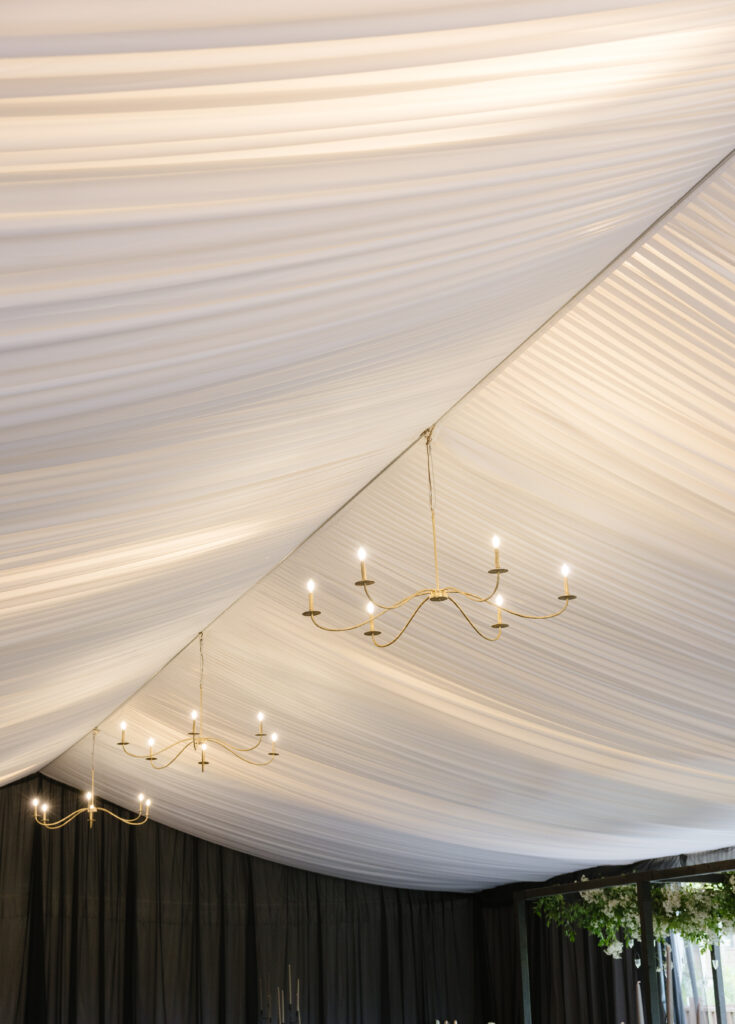 Elegant draped tent ceiling with modern gold chandeliers, creating a soft and romantic reception atmosphere. 