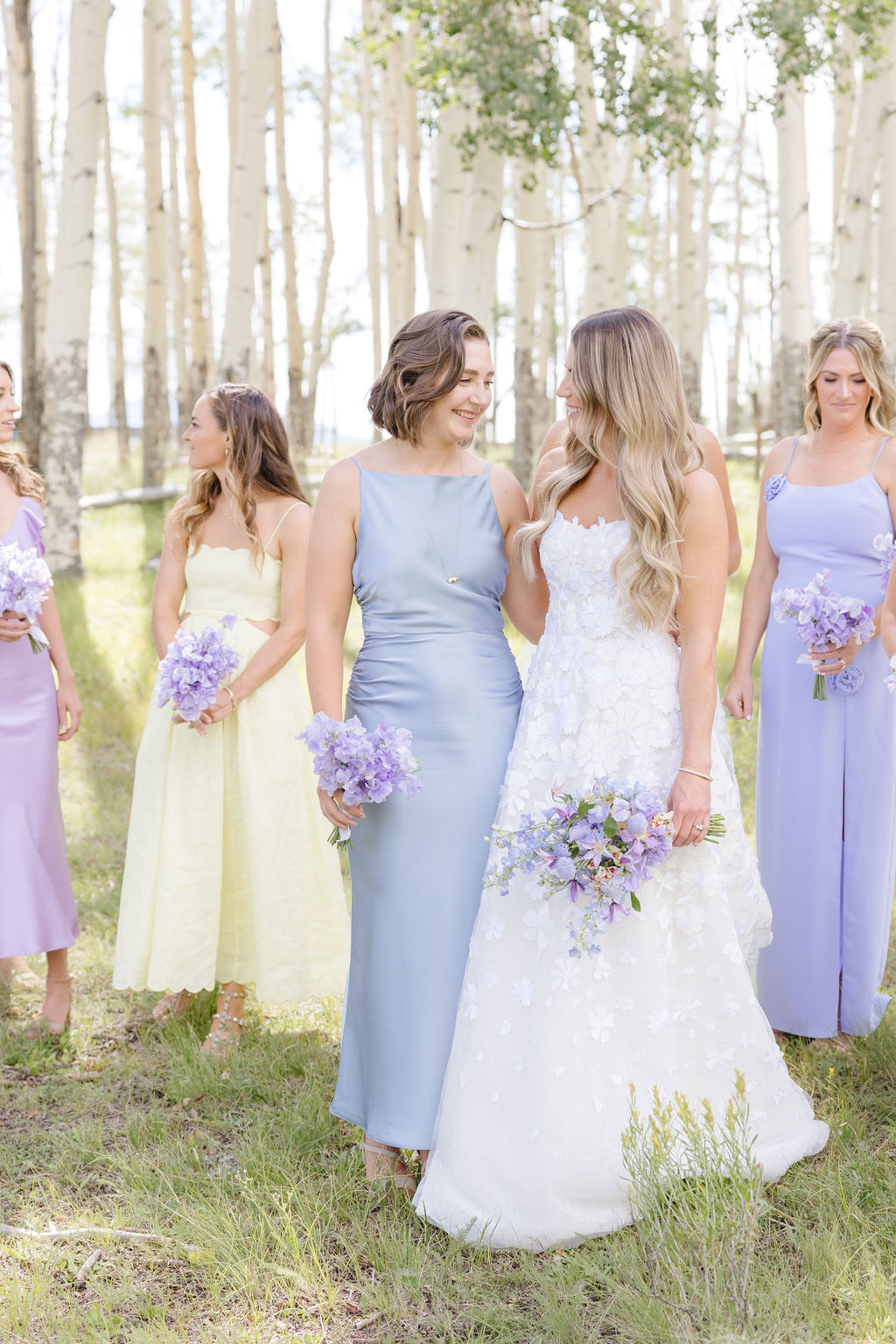 Bride and bridesmaids in spring colors walk through birch tree forest.