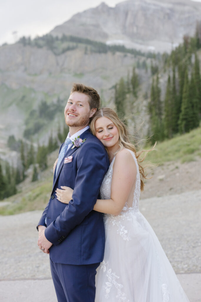 Bride hugs groom from behind with mountain views in the background.