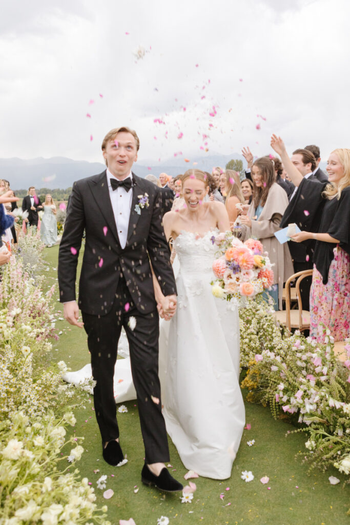 Bride and groom walk down aisle as guests throw flower petals. 