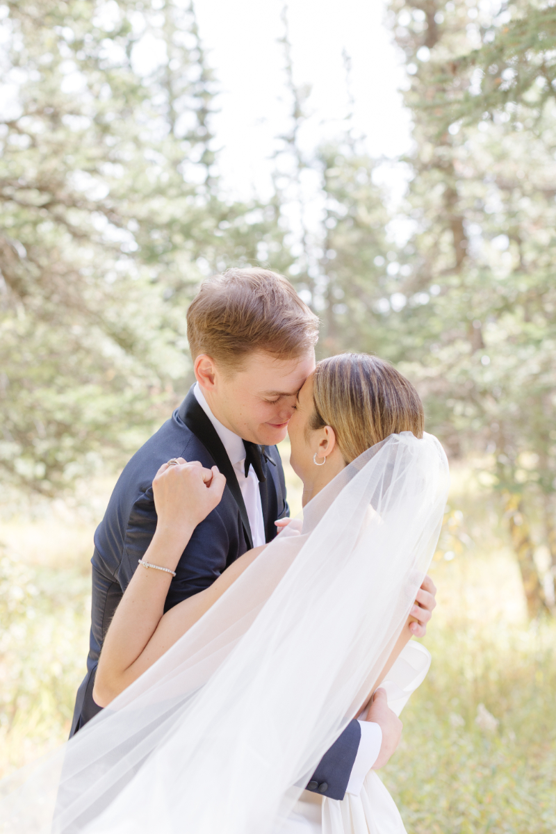 Bride and groom embrace, surrounded by pine trees.