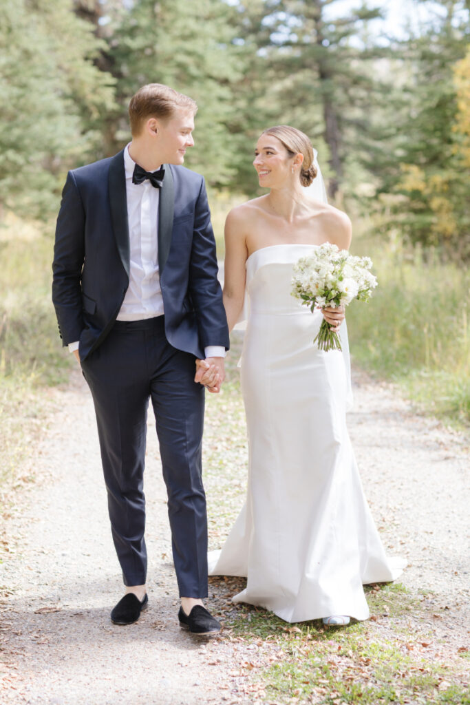 Bride and groom walk together on gravel road as their photographer captures portraits.