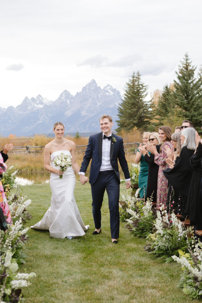 Bride and groom walk hand in hand as they exit their ceremony, towering views of the Tetons behind them, guests surrounding them cheering at this beautiful Moose Creek Ranch wedding.