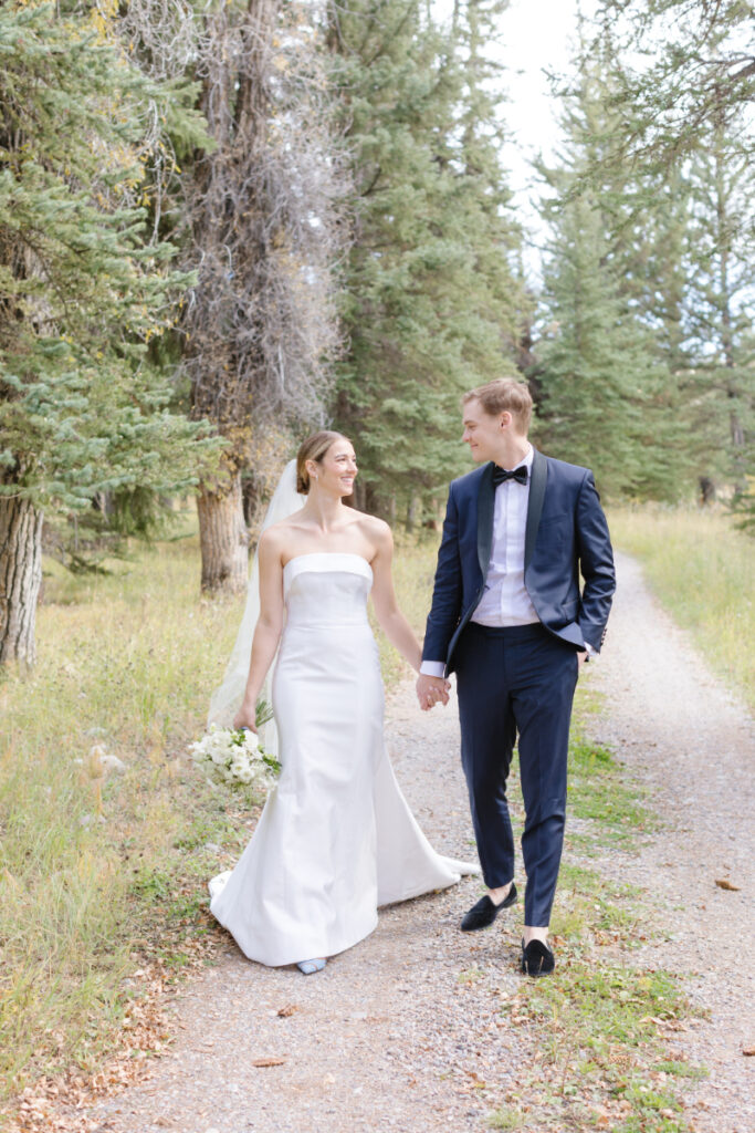 Bride and groom walk hand in hand, looking at each other, as they walk toward photographer on a dirt road in the Tetons for wedding portraits.