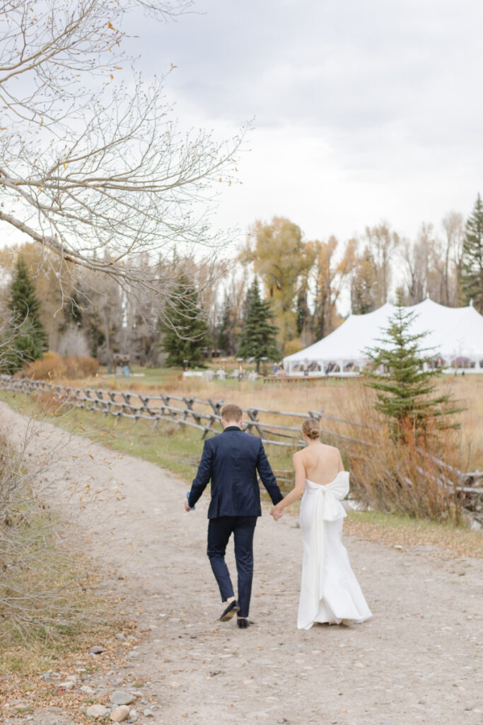 Bride and groom walk hand in hand on a gravel road leading them to their tented reception at their Moose Creek Ranch wedding.