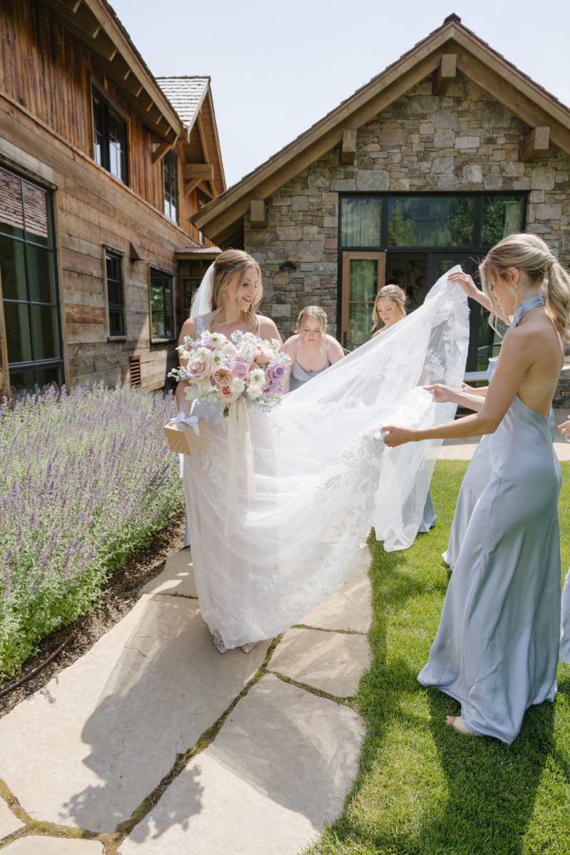 Bridesmaids help bride walk along sidewalk of destination wedding venue in Jackson Hole as they hold the train of her dress.
