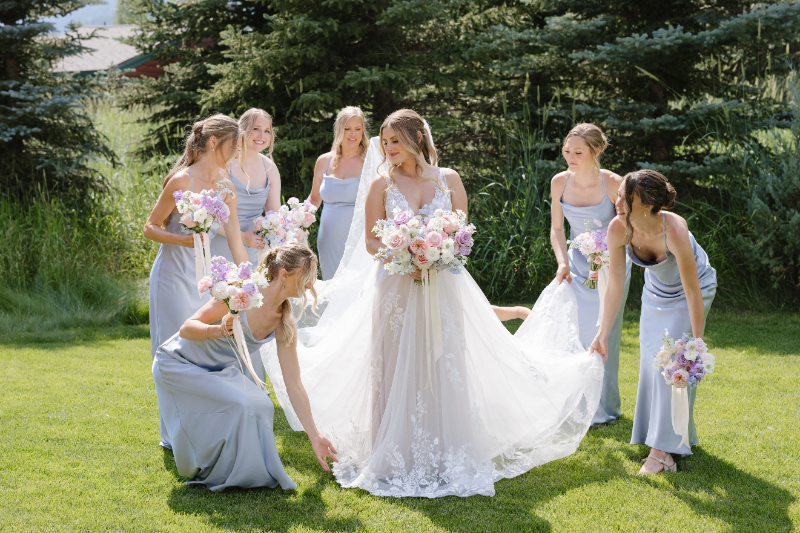 Bride stands on ground of Hotel Yellowstone at Jackson Hole as her bridesmaids surround her, fluffing her dress.