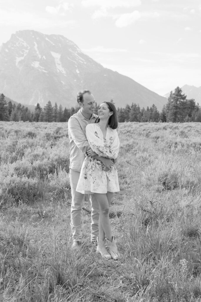 Black and white photo of couple. The man his hugging from behind and smiling at his wife-to-be.