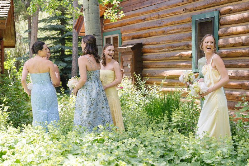 Bridesmaids in butter yellow and sky blue dresses walk in tall grassy area next to getting ready cabin.