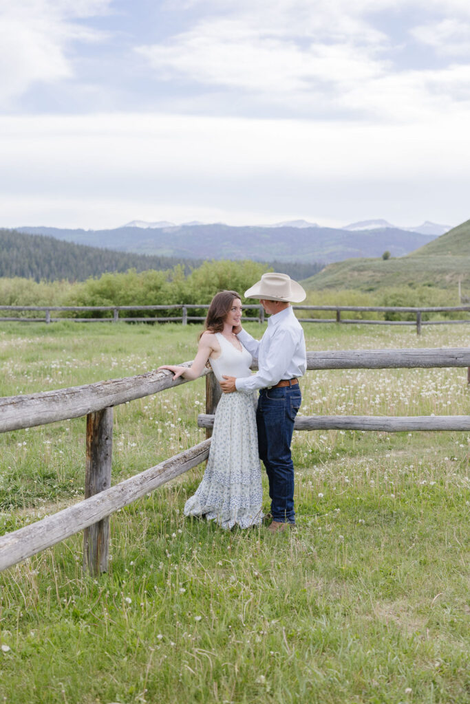 Couple standing by a wooden fence in a mountain meadow during romantic lake engagement photos