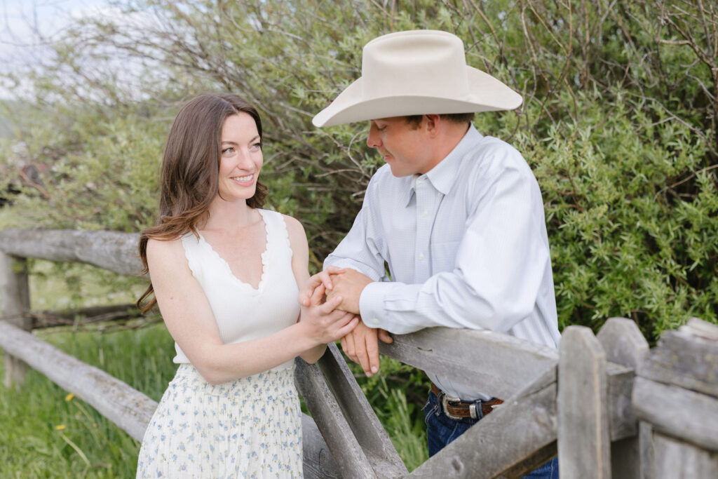 Couple holding hands and smiling at each other while leaning on a wooden fence in a grassy field