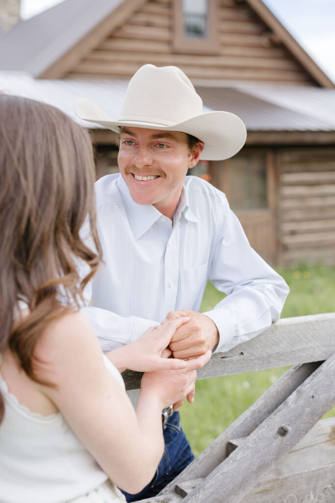 Man in a cowboy hat smiling at his partner during an engagement session near a rustic cabi