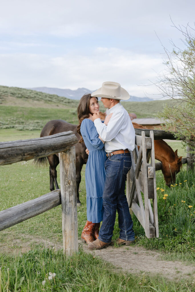 Couple standing by a wooden fence with horses nearby during rustic lake engagement photos in Jackson Hole
