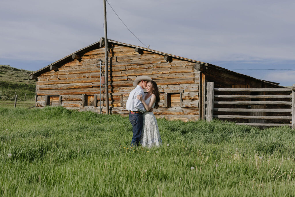Couple embracing in a grassy field near a cabin during golden hour lake engagement photos in the mountains