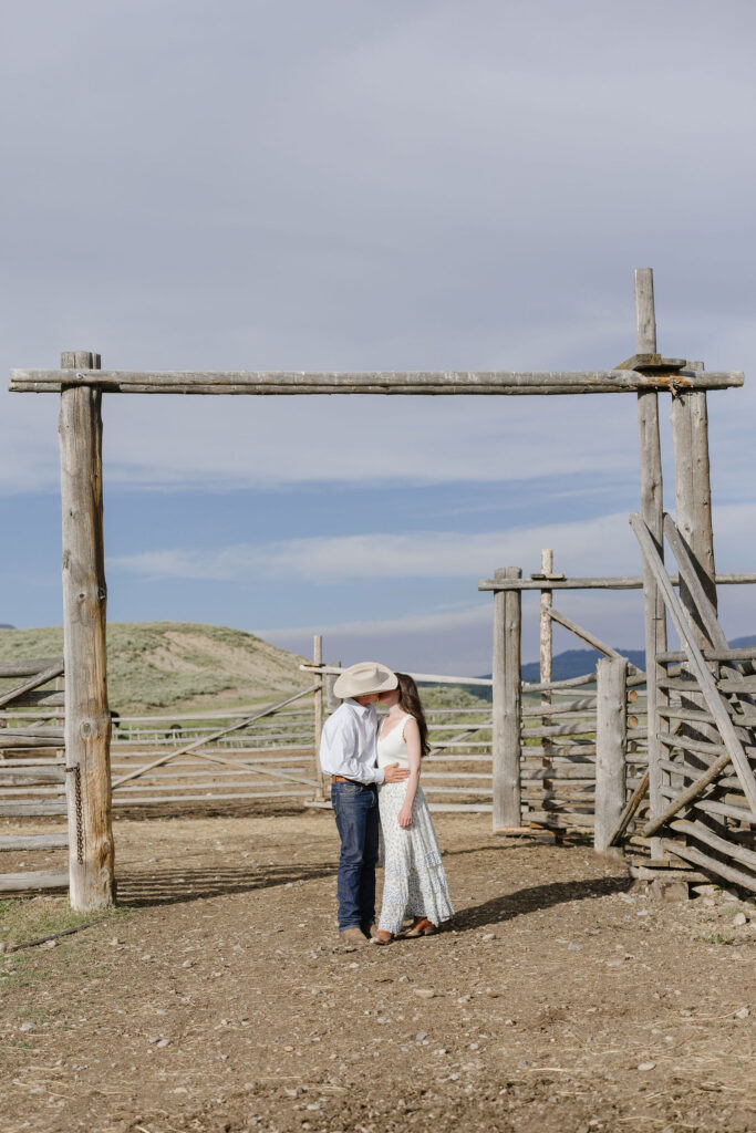Couple kissing beneath rustic wooden posts during sunlit lake engagement photos in a western setting