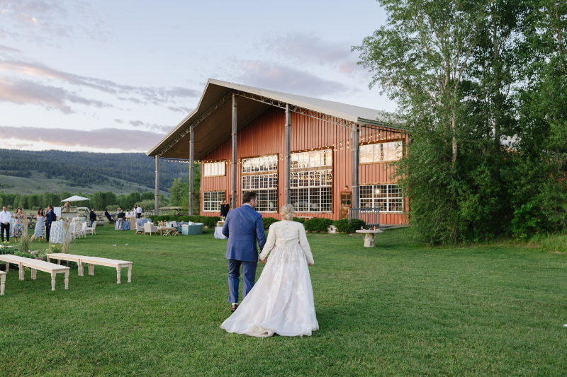 Bride and groom walking toward a rustic barn reception during a Western wedding in Jackson Hole with mountain ranch views at sunset.