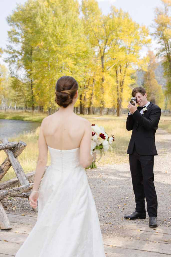 Groom taking a photo of the bride by a river with fall trees during a Jackson Hole wedding