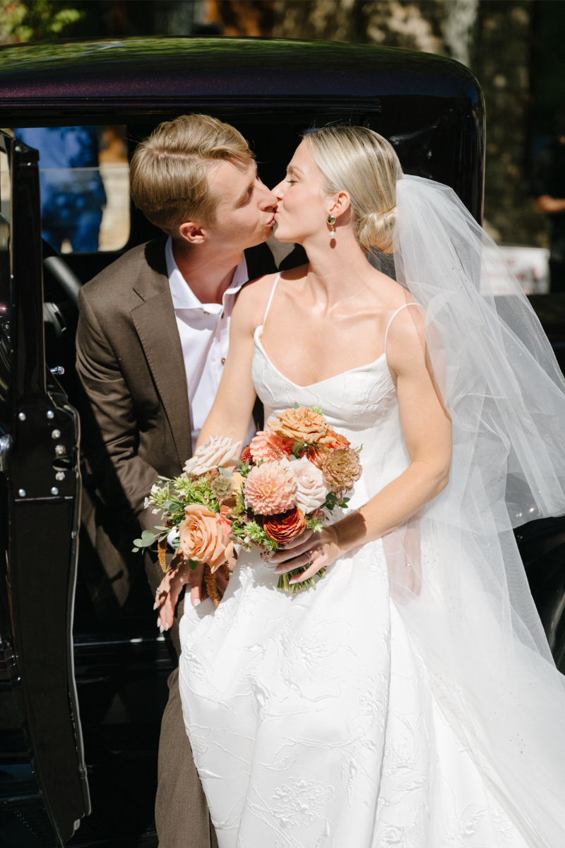 Bride and groom sharing a kiss beside a vintage getaway car after their wedding ceremony at Snake River Sporting Club