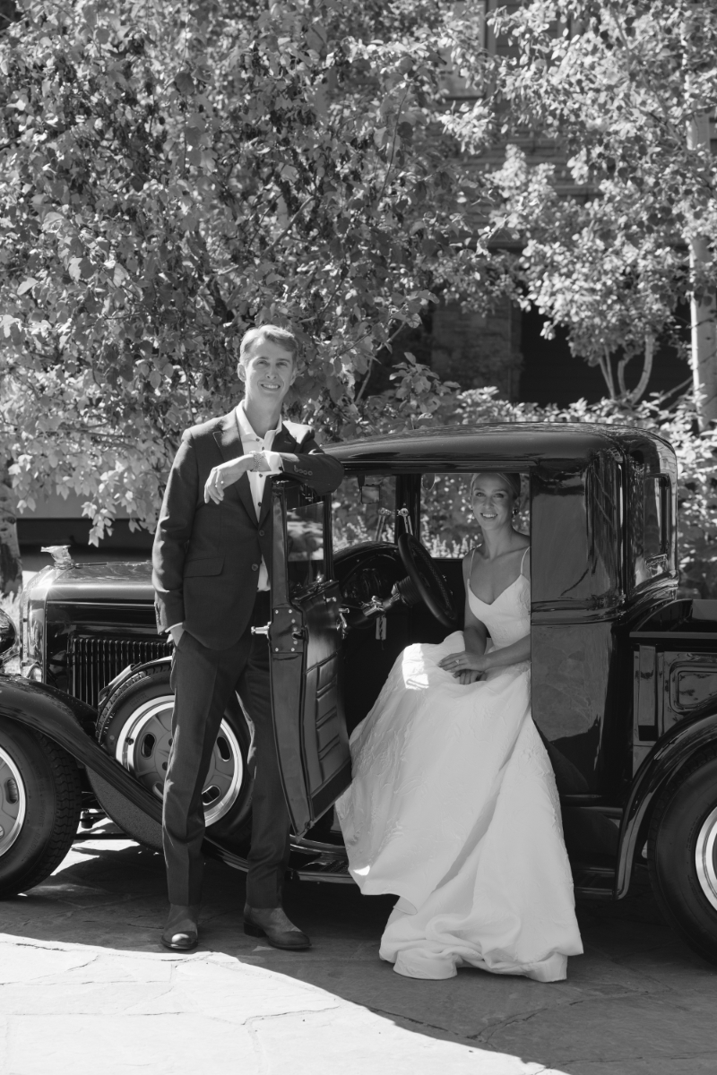 Bride sits in vintage getaway car at Snake River Sporting Club as groom leans against the door before they leave for their ceremony.