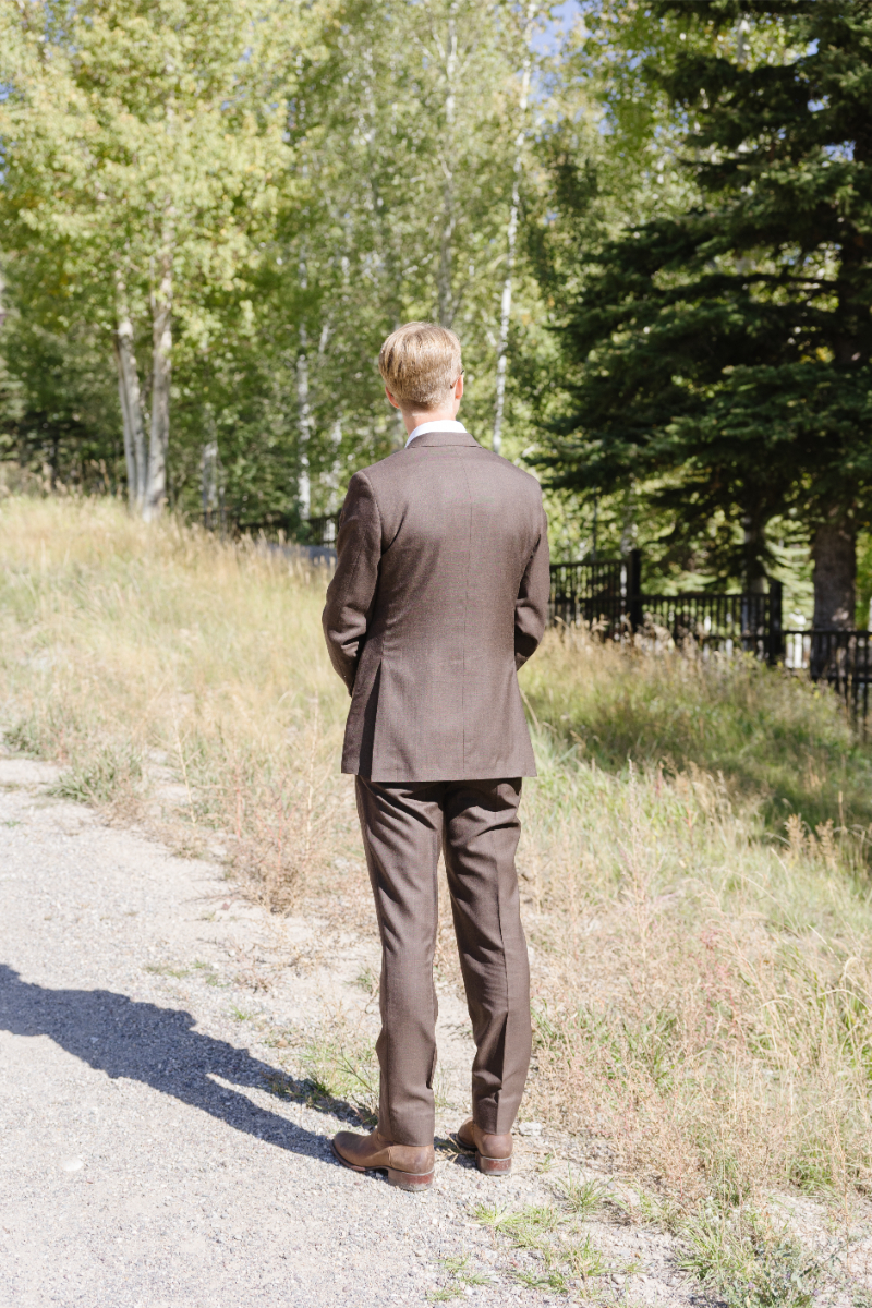 Groom standing in brown tux facing away from bride before their first look.