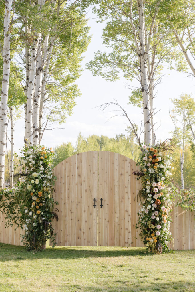 Wooden ceremony entrance framed by floral installations beneath towering aspen trees at a Jackson Hole wedding venue.