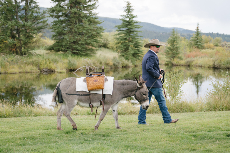 A man in western attire leads a pack donkey carrying a picnic basket beside a pond at a Jackson Hole wedding venue.