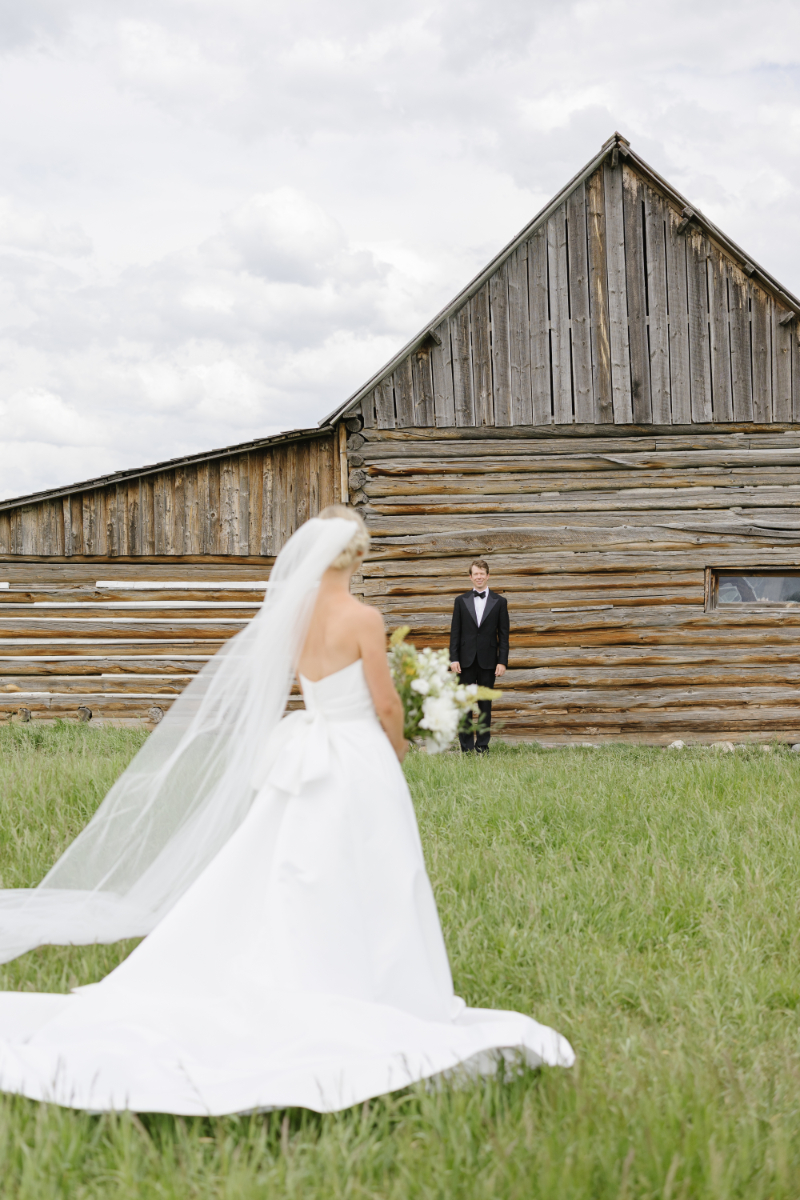 Bride approaches groom for their first look outside a rustic log barn during a Jackson Hole wedding.