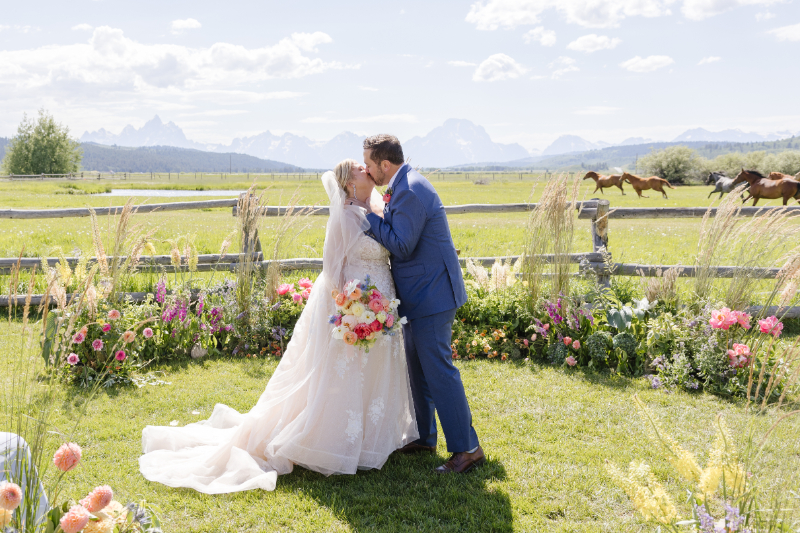 Bride and groom kissing beside colorful flowers with horses and the Teton landscape behind them